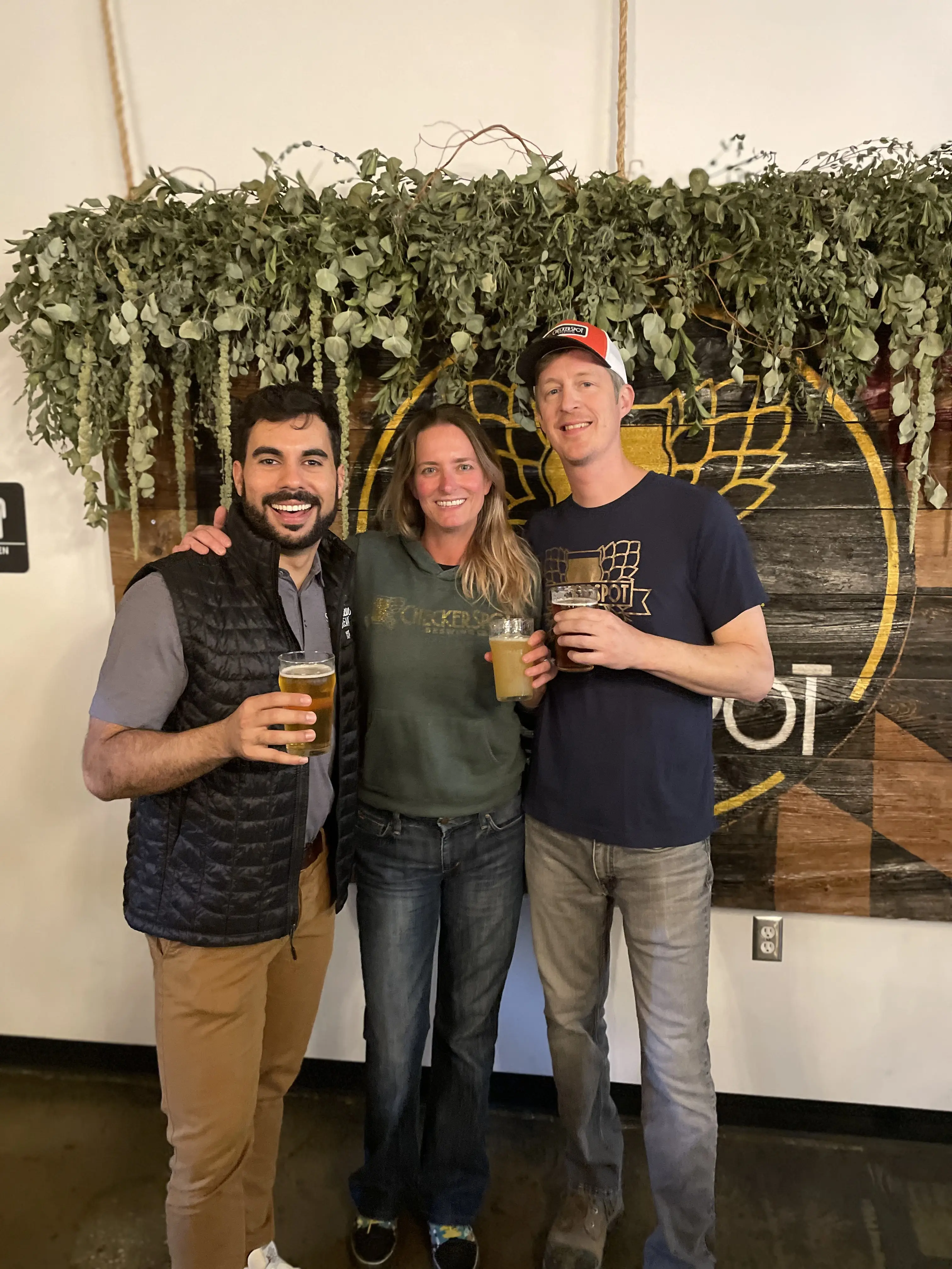 The Checkerspot crew raising a glass in front of the brewery's logo wall