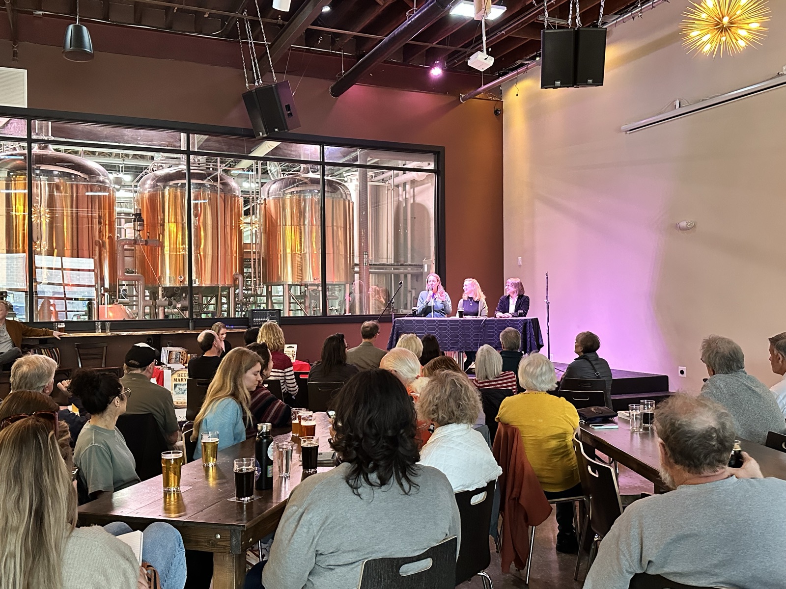 A seated crowd at a panel talk with the copper brewhouse tanks glowing behind glass
